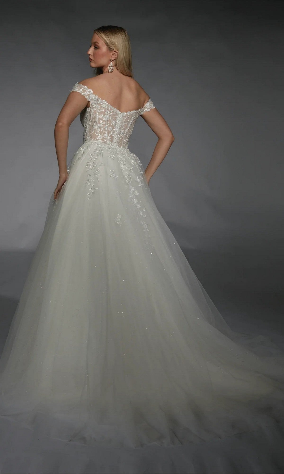 A woman stands facing away against a dark background, wearing the Alyce 7129 long white dress featuring an off-the-shoulder, ivory ball gown design with a lace corset bodice and floral details.